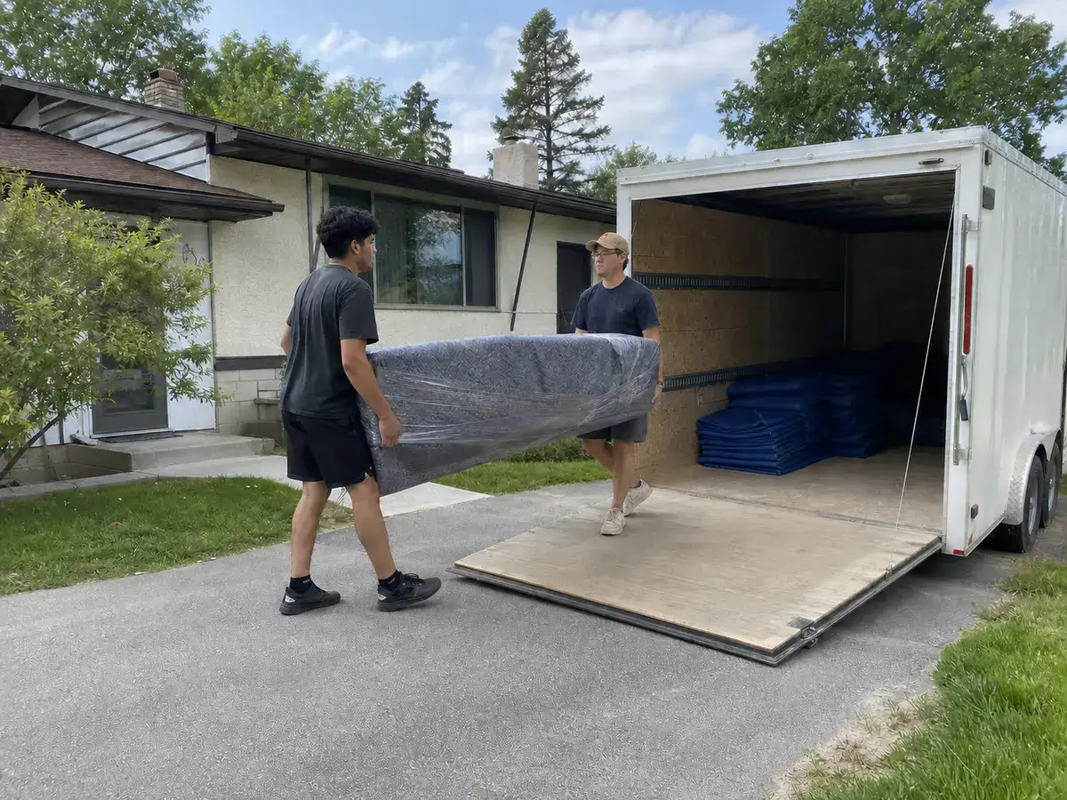 Two Winnipeg movers carrying a wrapped sofa toward a moving trailer as one coordinated crew